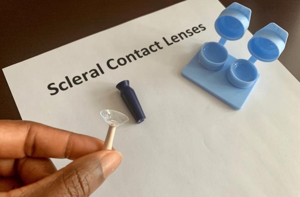Close-up of a person's hand holding an insertion tool with a scleral contact lens above a paper that reads "Scleral Contact Lenses" with a contact lens case resting on it