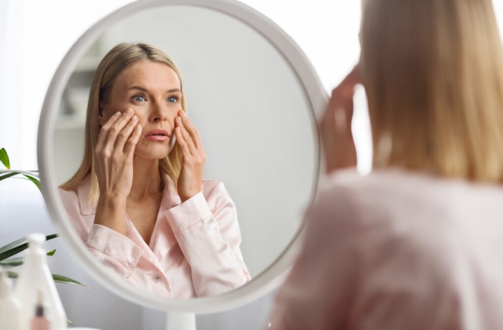 A person looks at their face in the mirror, examining the skin under their eyes for signs of dryness.