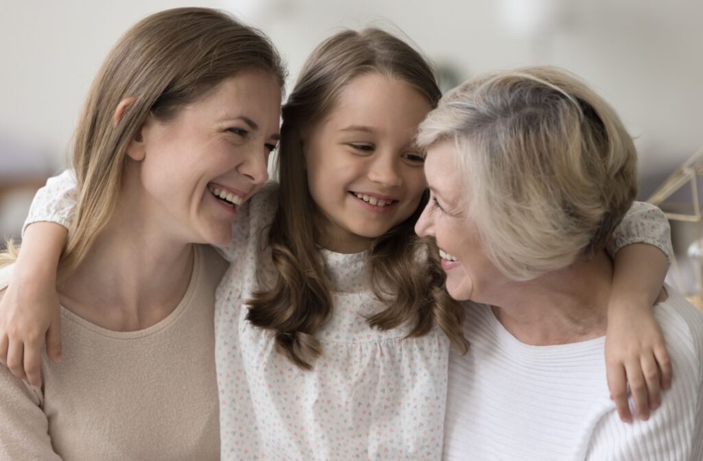 Three intergenerational family members in white and beige clothing hold each other in a hug, with the youngest in the middle