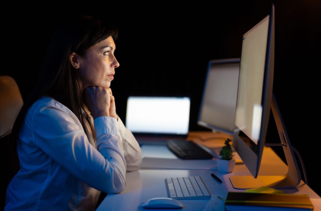 A person sits in a dark room facing a bright computer screen in front of them.