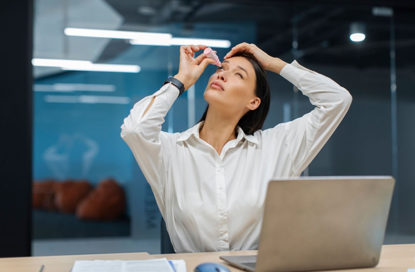 A person applies eye drops while sitting at a desk with an open laptop, looking upward and pulling down their lower eyelid.